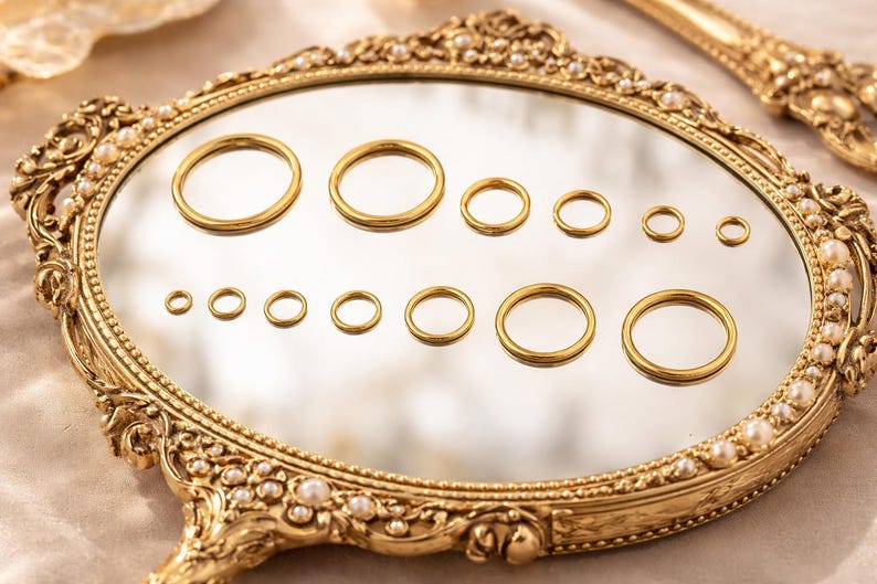 May include: A collection of twelve gold-toned metal rings of varying diameters arranged in two rows on a reflective surface. The rings are displayed on an ornate, oval-shaped gold frame decorated with small white pearls along its edge.