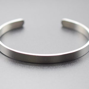 K&ouml;nnte beinhalten: Ein silberfarbenes Armreif-Armband. Das Armband hat ein schlichtes, minimalistisches Design mit einem flachen, rechteckigen Band von etwa 0,6 cm Breite und abgerundeten Kanten. Das offene Armband besteht aus gl&auml;nzendem Metall.