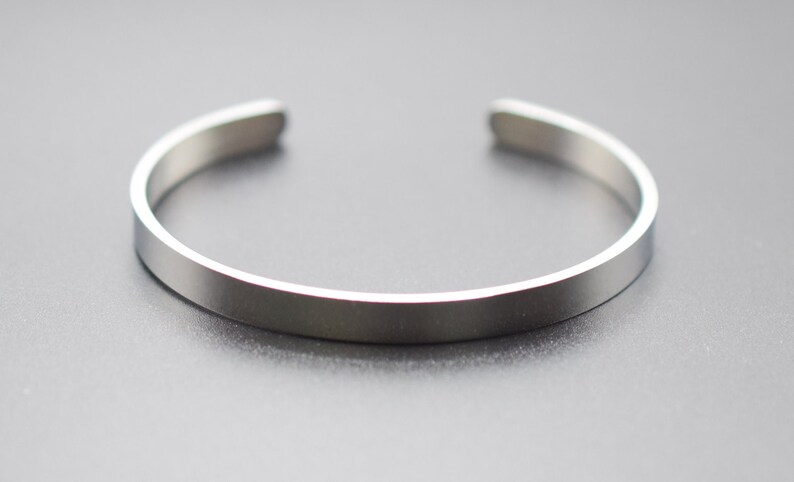 K&ouml;nnte beinhalten: Ein silberfarbenes Armreif-Armband. Das Armband hat ein schlichtes, minimalistisches Design mit einem flachen, rechteckigen Band von etwa 0,6 cm Breite und abgerundeten Kanten. Das offene Armband besteht aus gl&auml;nzendem Metall.