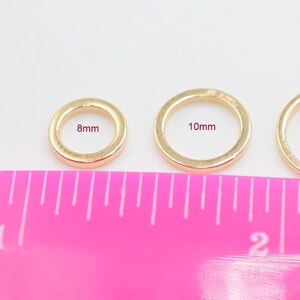 May include: Three gold-toned circular rings arranged in a row, labeled with their respective diameters of 8mm, 10mm, and 12mm. A bright pink ruler is positioned directly below the rings, showing measurements in inches for scale and size reference.