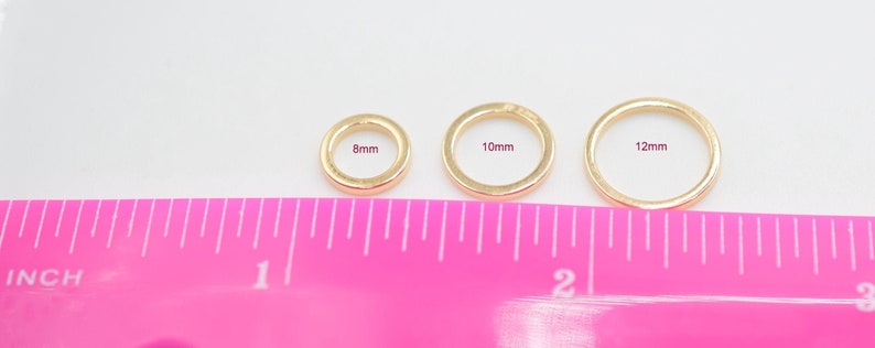 May include: Three gold-toned circular rings arranged in a row, labeled with their respective diameters of 8mm, 10mm, and 12mm. A bright pink ruler is positioned directly below the rings, showing measurements in inches for scale and size reference.