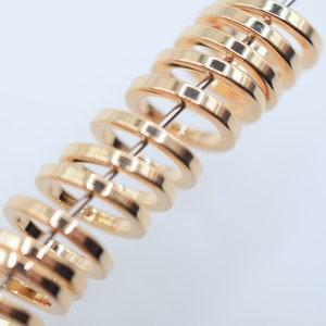 May include: A series of small, circular, gold-toned metal rings strung together on a thin, straight wire. Each ring features a smooth, polished surface with a reflective finish, arranged in a repeating, evenly spaced vertical alignment.
