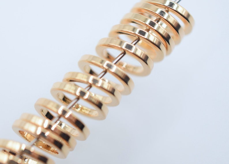 May include: A series of small, circular, gold-toned metal rings strung together on a thin, straight wire. Each ring features a smooth, polished surface with a reflective finish, arranged in a repeating, evenly spaced vertical alignment.