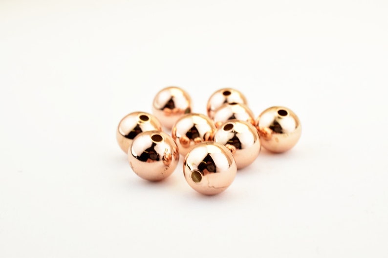 Peut inclure: Un groupe de dix perles rondes en or rose avec un trou au centre. Les perles sont dispos&eacute;es en un groupe l&acirc;che sur un fond blanc.