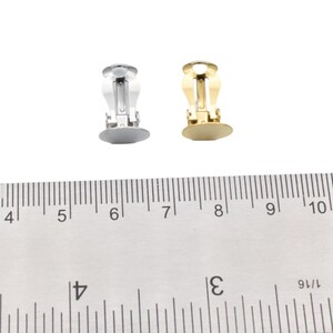 May include: Two pairs of earring clips, one silver and one gold, are shown on a white surface next to a ruler. The ruler is marked in inches.