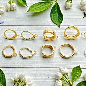 Puede incluir: Varios modelos de cierres de pendientes dorados, incluyendo dise&ntilde;os de palanca y aro, se disponen sobre una superficie de madera blanca. La imagen est&aacute; decorada con flores blancas y hojas verdes, creando una presentaci&oacute;n visualmente atractiva.