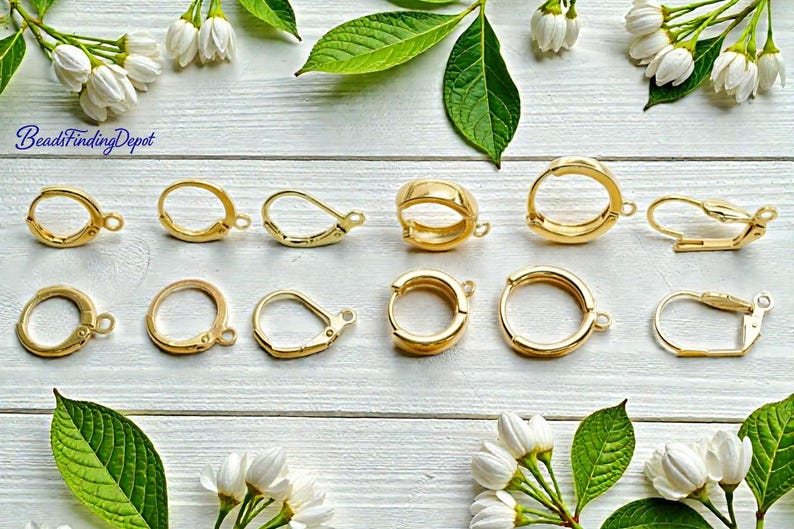 Puede incluir: Varios modelos de cierres de pendientes dorados, incluyendo dise&ntilde;os de palanca y aro, se disponen sobre una superficie de madera blanca. La imagen est&aacute; decorada con flores blancas y hojas verdes, creando una presentaci&oacute;n visualmente atractiva.