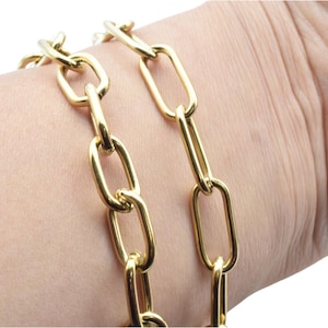 May include: A gold-colored chain bracelet with elongated oval links. The bracelet is made of a shiny, metallic material and is worn on a wrist. The links are connected in a repeating pattern, creating a bold and modern look.