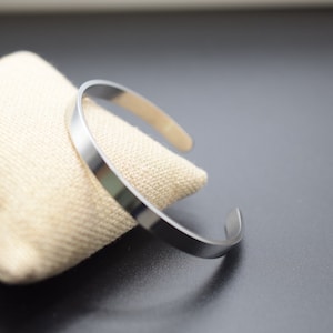K&ouml;nnte beinhalten: Ein silberfarbenes Armreif-Armband mit schlichtem, minimalistischem Design. Das offene Armband liegt auf einem beigefarbenen Stoff. Der Hintergrund ist eine dunkelgraue Oberfl&auml;che.