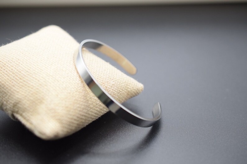 K&ouml;nnte beinhalten: Ein silberfarbenes Armreif-Armband mit schlichtem, minimalistischem Design. Das offene Armband liegt auf einem beigefarbenen Stoff. Der Hintergrund ist eine dunkelgraue Oberfl&auml;che.