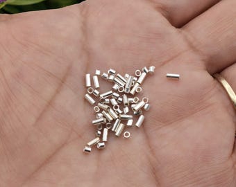 May include: A collection of small, cylindrical silver-colored metal beads scattered across a palm. Each bead features a hollow center and a smooth, reflective surface. The beads are uniform in shape and size, appearing as tiny metallic tubes.