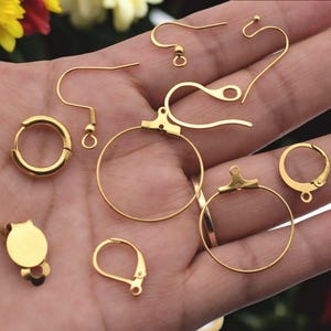 May include: An assortment of gold-coloured earring findings, including different styles of hooks, hoops, and clasps. The components are displayed against a soft, neutral background, showcasing the different designs and sizes available for jewellery making.