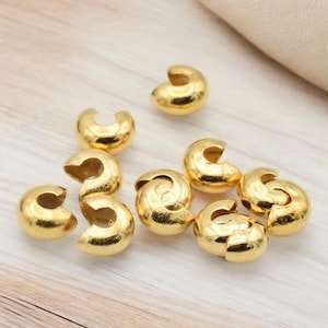 May include: A group of ten gold-colored metal crimp beads. The beads are round and have a small opening on one side. They are used to secure the ends of cords or strings.