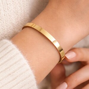 K&ouml;nnte beinhalten: Ein goldfarbenes, minimalistisches Armband. Das polierte, flache Band umschlie&szlig;t das Handgelenk. Das Armband wird an einem hellh&auml;utigen Arm getragen, kombiniert mit einem cremefarbenen Pullover. Das schlichte Design macht es zu einem vielseitigen Accessoire.