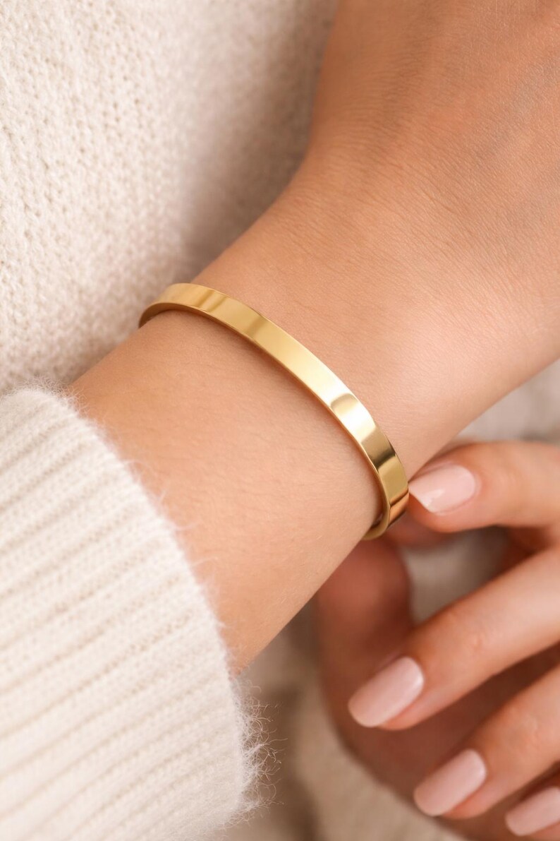 K&ouml;nnte beinhalten: Ein goldfarbenes, minimalistisches Armband. Das polierte, flache Band umschlie&szlig;t das Handgelenk. Das Armband wird an einem hellh&auml;utigen Arm getragen, kombiniert mit einem cremefarbenen Pullover. Das schlichte Design macht es zu einem vielseitigen Accessoire.