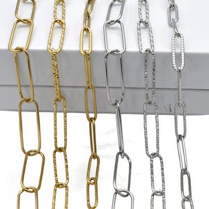 May include: Close-up of several gold and silver chain necklaces with elongated oval links. The chains have different textures, including smooth and hammered finishes. The necklaces are displayed against a white background, showcasing their varying metallic tones.