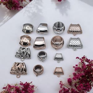 May include: A collection of jewelry findings in various metallic finishes, including silver, gold, and rose gold. The assortment features different shapes, such as rounded, rectangular, and decorative designs with floral and textured patterns. Each finding has a loop for attaching to jewelry.