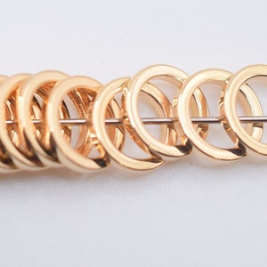 May include: A row of polished gold-tone metal jump rings threaded onto a thin silver-colored wire. The circular rings are uniform in size and shape, featuring a smooth, reflective surface that catches the light, arranged in a horizontal line.