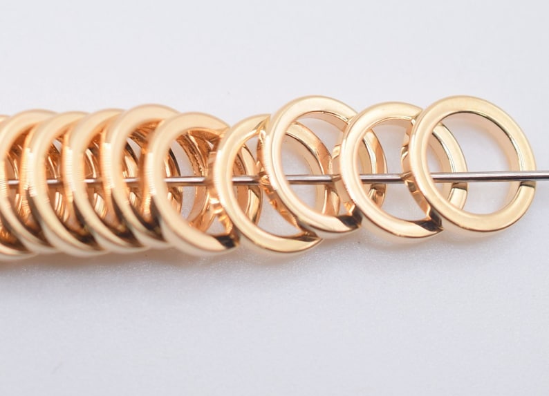 May include: A row of polished gold-tone metal jump rings threaded onto a thin silver-colored wire. The circular rings are uniform in size and shape, featuring a smooth, reflective surface that catches the light, arranged in a horizontal line.