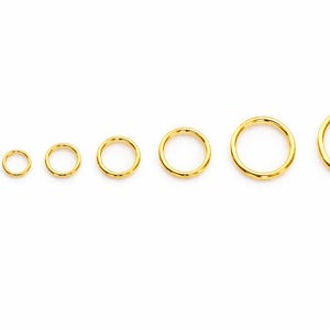 May include: A set of eight circular gold-toned metal jump rings arranged in a horizontal row from smallest to largest. Each ring features a smooth, polished surface and a continuous, seamless design, showcasing a consistent metallic yellow finish.