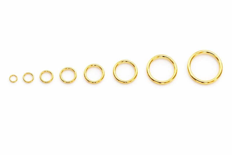 May include: A set of eight circular gold-toned metal jump rings arranged in a horizontal row from smallest to largest. Each ring features a smooth, polished surface and a continuous, seamless design, showcasing a consistent metallic yellow finish.