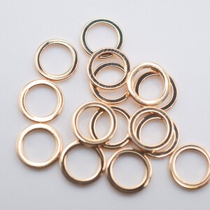 May include: A collection of small, circular, metallic jump rings with a polished rose gold finish. These open-ended connectors are arranged in a loose, overlapping cluster, showcasing their smooth, uniform, and reflective circular design.