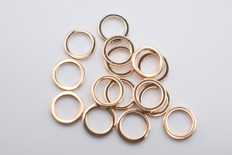 May include: A collection of small, circular, metallic jump rings with a polished rose gold finish. These open-ended connectors are arranged in a loose, overlapping cluster, showcasing their smooth, uniform, and reflective circular design.