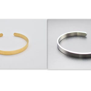 K&ouml;nnte beinhalten: Zwei offene Armreifen. Einer ist goldfarben, der andere silberfarben. Die Armreifen sind schlicht, mit einem flachen, abgerundeten Band. Der goldene Armreif befindet sich auf wei&szlig;em Hintergrund, der silberne auf dunklem Grau.
