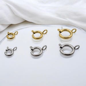 May include: A collection of six circular spring ring clasps arranged in two rows. The top row features three gold-toned metal clasps in varying sizes, while the bottom row displays three silver-toned metal clasps in matching sizes, each with a small loop.