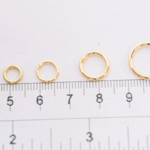 May include: A series of eight gold-colored metal jump rings arranged in increasing order of size, positioned along a ruler that shows measurements in both inches and centimeters to provide a clear scale for the diameter of each individual ring.