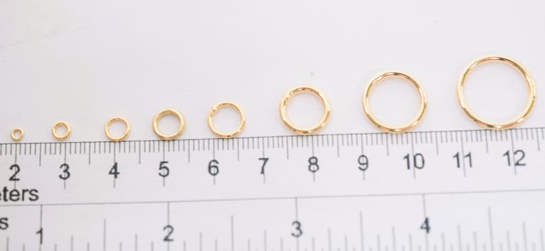 May include: A series of eight gold-colored metal jump rings arranged in increasing order of size, positioned along a ruler that shows measurements in both inches and centimeters to provide a clear scale for the diameter of each individual ring.