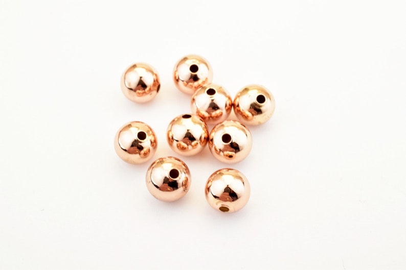 Peut inclure: Dix perles de couleur or rose, chacune avec un petit trou au centre. Les perles sont &eacute;parpill&eacute;es sur une surface blanche.