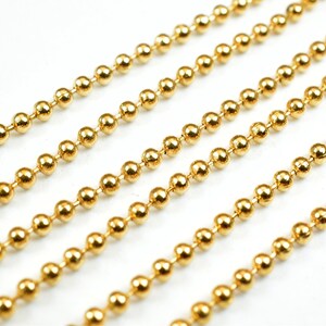 May include: A close-up of a gold ball chain. The chain is made of small, round beads that are linked together. The chain is lying flat on a white surface.