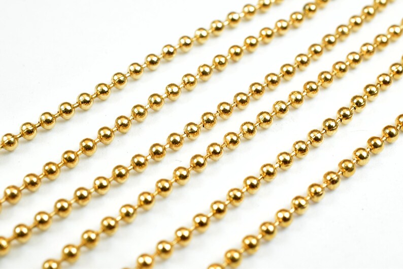 May include: A close-up of a gold ball chain. The chain is made of small, round beads that are linked together. The chain is lying flat on a white surface.