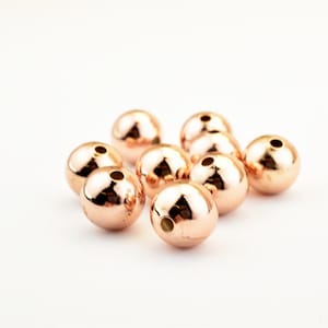 Peut inclure: Un groupe de dix perles rondes en or rose avec un trou au centre. Les perles sont dispos&eacute;es en un groupe l&acirc;che sur un fond blanc.