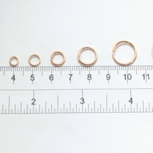 May include: A series of eight rose gold jump rings arranged in increasing order of size above a ruler. The ruler displays both metric and imperial measurements, with the largest ring measuring approximately 0.5 inches in diameter.
