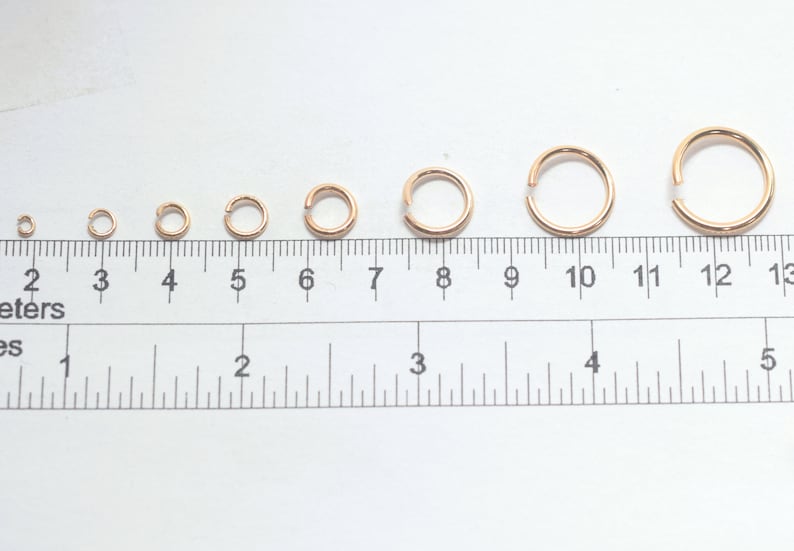 May include: A series of eight rose gold jump rings arranged in increasing order of size above a ruler. The ruler displays both metric and imperial measurements, with the largest ring measuring approximately 0.5 inches in diameter.