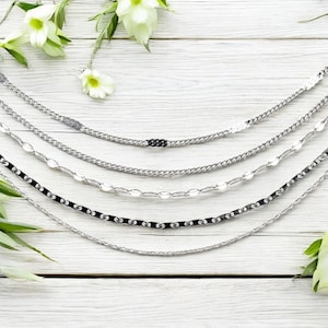 May include: An assortment of silver necklaces with various chain designs, including curb, oval, and twisted patterns. The necklaces are displayed on a white wooden surface, with white flowers and green leaves in the background.