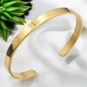 K&ouml;nnte beinhalten: Ein goldfarbenes Armreif-Armband mit flachem, minimalistischem Design. Das offene Armband besteht aus gl&auml;nzendem, glattem Metall. Das Armband wird auf einer wei&szlig;en Oberfl&auml;che mit einer gr&uuml;nen Pflanze im Hintergrund gezeigt. Ein schlichtes, elegantes Schmuckst&uuml;ck.