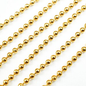 May include: A close-up of gold ball chain. The chain is made of small, round beads that are linked together. The chain is lying flat on a white surface.
