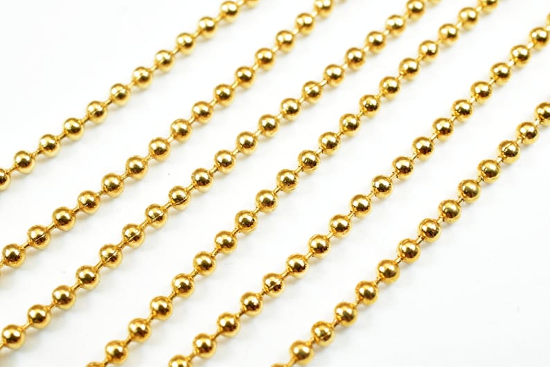 May include: A close-up of gold ball chain. The chain is made of small, round beads that are linked together. The chain is lying flat on a white surface.