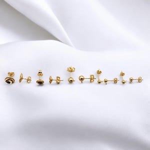 May include: A collection of gold-colored stud earrings in various designs, including ball, bar, and geometric shapes. The earrings are displayed on a white, textured surface, showcasing their polished finish and diverse styles.