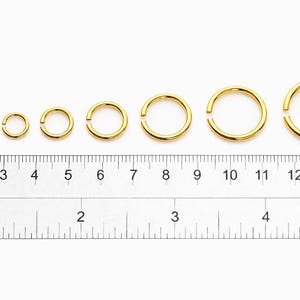 May include: A set of eight gold-colored metal jump rings arranged in increasing order of size above a ruler showing measurements in both centimeters and inches, with the smallest ring at 0.25 inches and the largest at 0.5 inches in diameter.