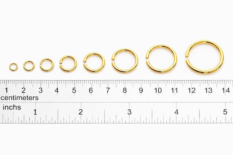 May include: A set of eight gold-colored metal jump rings arranged in increasing order of size above a ruler showing measurements in both centimeters and inches, with the smallest ring at 0.25 inches and the largest at 0.5 inches in diameter.