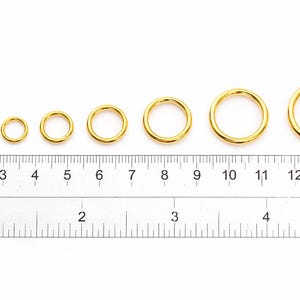 May include: Eight gold-colored metal jump rings arranged in increasing order of size, positioned above a ruler showing both centimeters and inches. The rings range from approximately 0.25 inches to 1 inch in diameter as measured by the ruler.