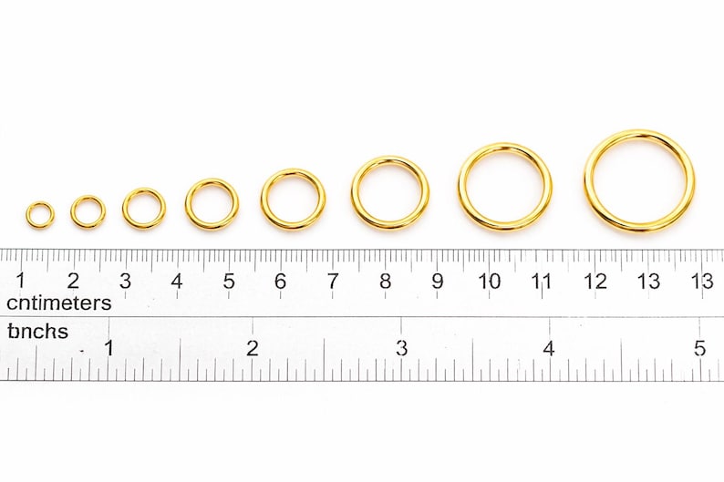 May include: Eight gold-colored metal jump rings arranged in increasing order of size, positioned above a ruler showing both centimeters and inches. The rings range from approximately 0.25 inches to 1 inch in diameter as measured by the ruler.