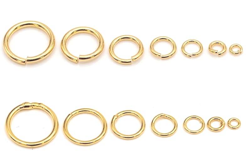 May include: A collection of fourteen gold-toned metal jump rings arranged in two horizontal rows. The rings are displayed in seven graduated sizes, ranging from large to small, each featuring a single split opening for jewelry assembly.