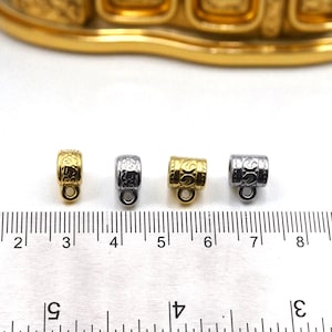 May include: Four small metal jewelry components. Two are gold-coloured, and two are silver-coloured. Each has a decorative pattern and a small loop at the bottom. A ruler is in the foreground, with a gold-coloured object in the background.