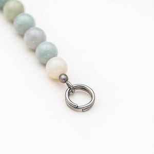 May include: A close-up of a beaded necklace with a silver clasp. The necklace features a strand of round, pastel-colored beads in shades of green, blue, and white. The clasp is a silver ring.