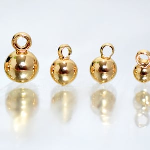 May include: A set of six polished gold-toned spherical charms arranged in a row, each featuring a small circular loop at the top for attachment. The charms vary in size from largest to smallest, displaying a smooth, reflective metallic surface.